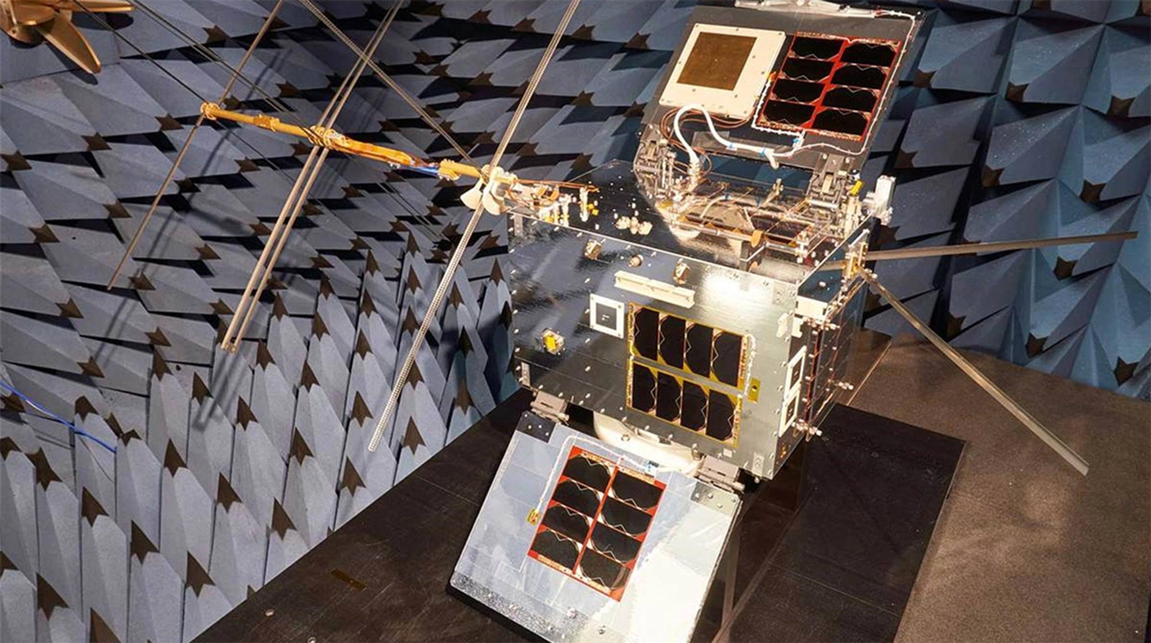 A compact satellite with multiple deployed antennas and solar panels is displayed in an anechoic chamber lined with pyramid-shaped blue absorbers. The satellite is positioned on a dark platform, with visible wiring and components exposed for testing or demonstration.