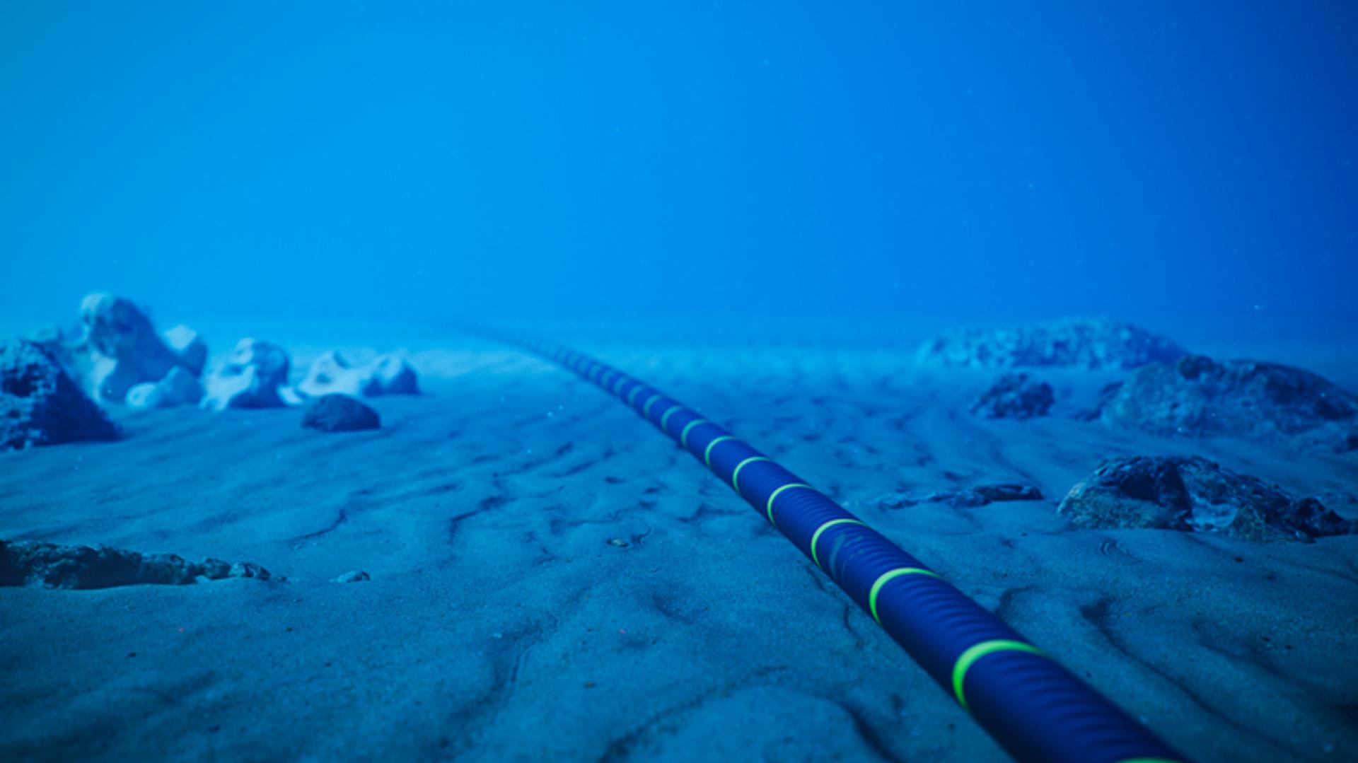 A submarine fiber optic cable lies on the ocean floor, stretching into the distance. The cable is black with bright green rings and is surrounded by sand and scattered rocks under deep blue water.