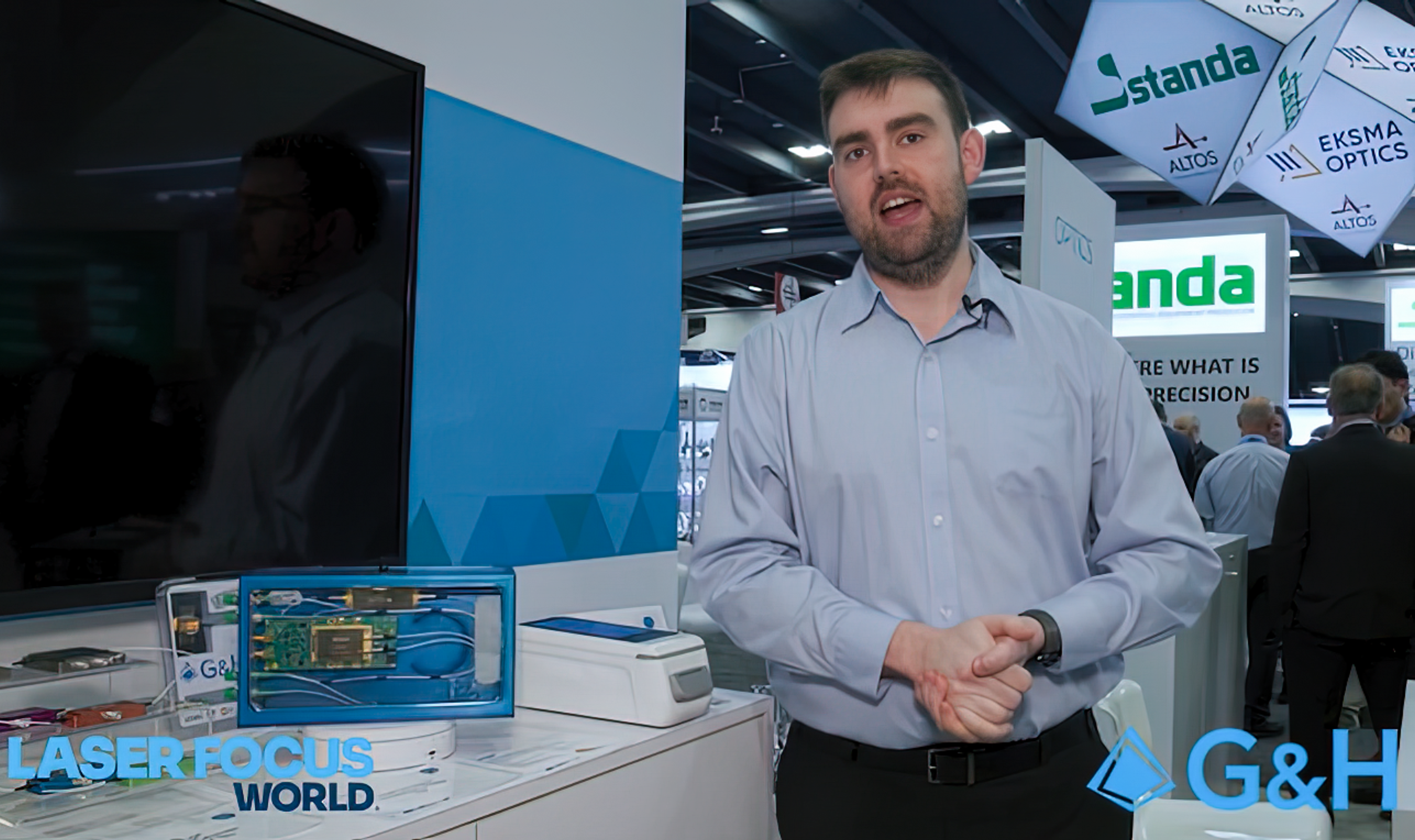 G&H representative speaking on-camera at a Photonics West trade show booth, with product display and Laser Focus World and G&H logos visible on screen.