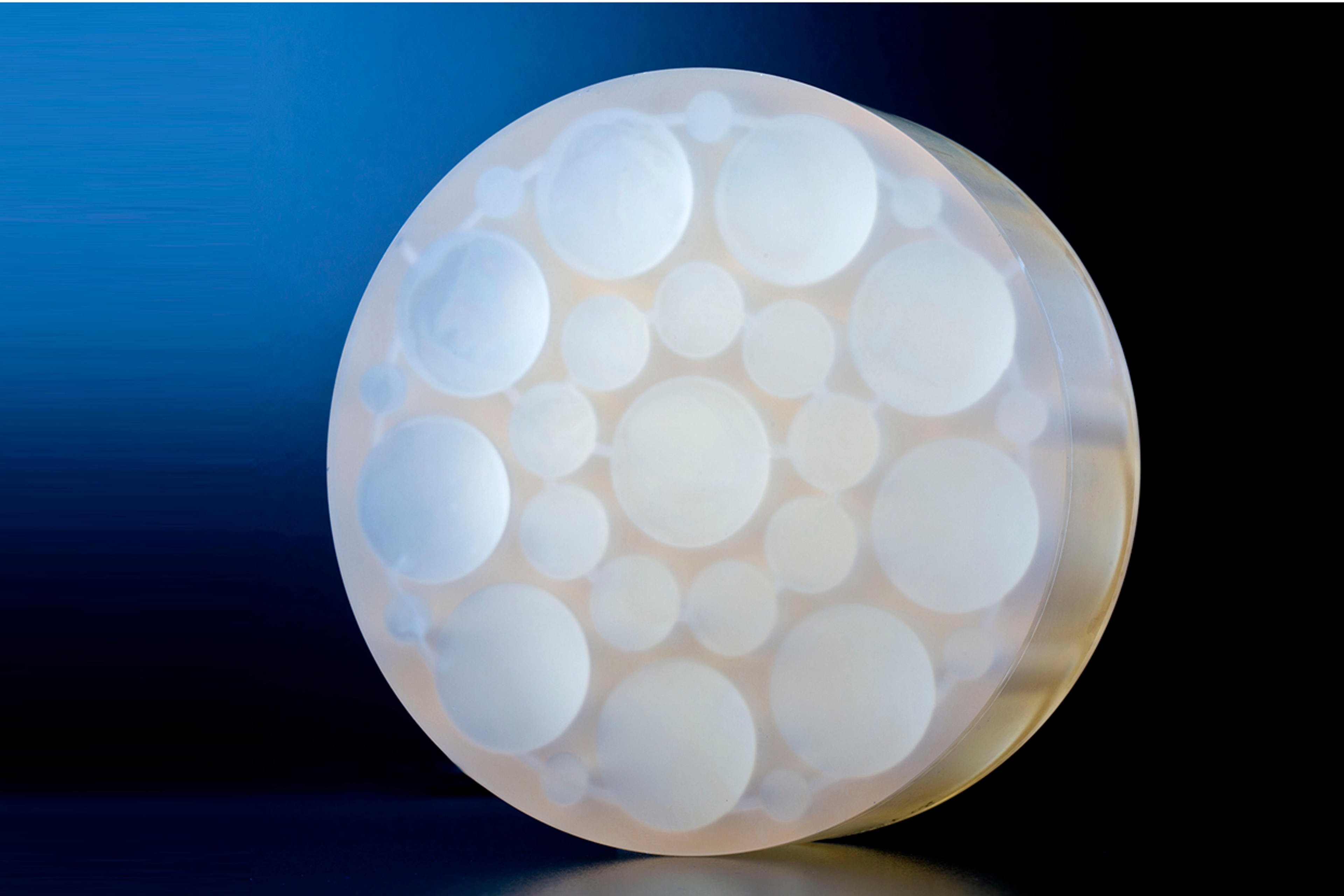 Hy-Per Bond™ - A close-up of a transparent circular optical component featuring multiple internal convex lens elements of varying sizes arranged in a honeycomb-like pattern, placed against a dark blue and black background.