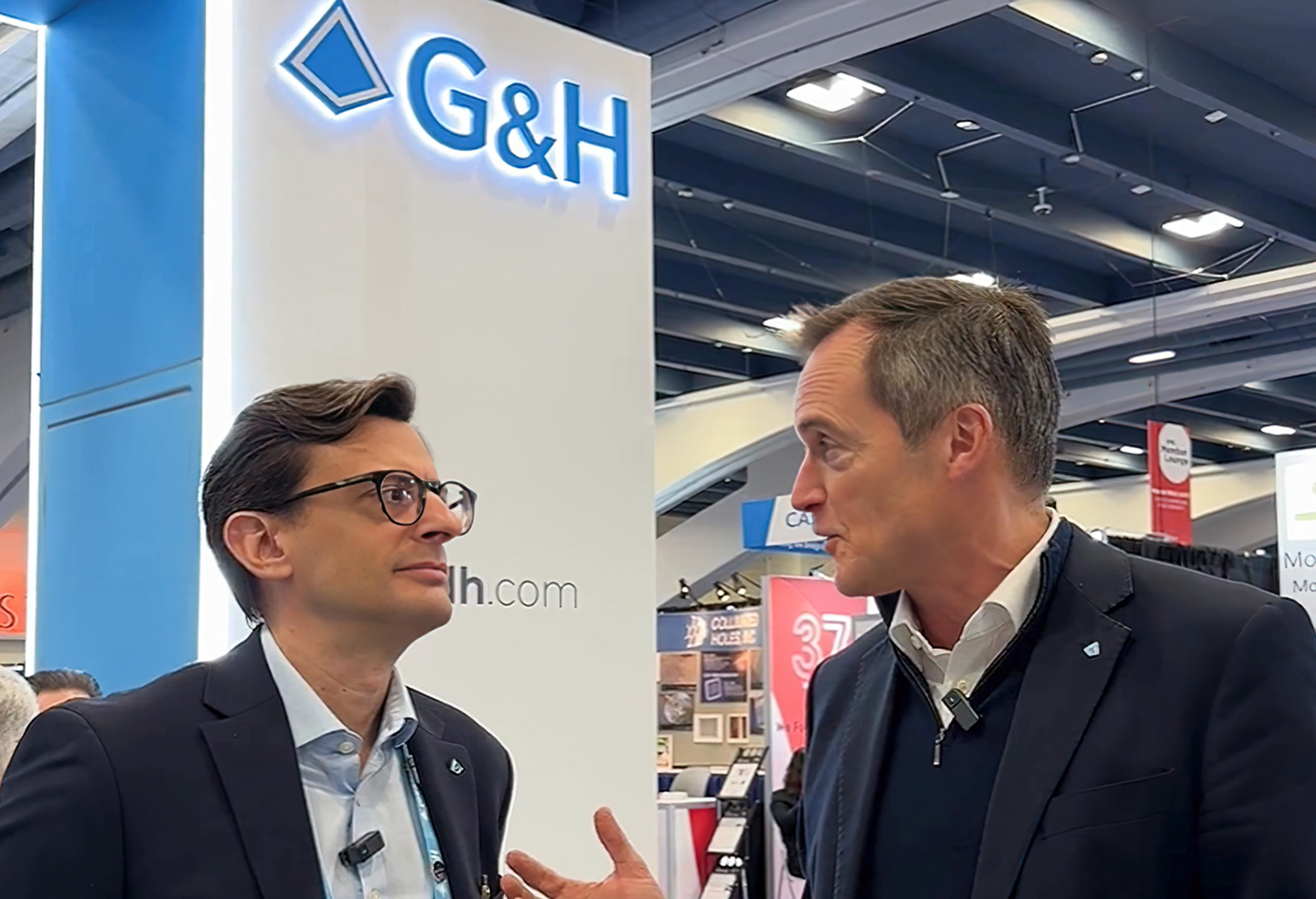 Charlie Peppiatt and Stratos Kehayas in conversation at the G&H Photonics West 2026 trade show stand, standing in front of a G&H exhibition booth with the illuminated G&H logo visible, discussing photonics and technology in a busy exhibition hall.