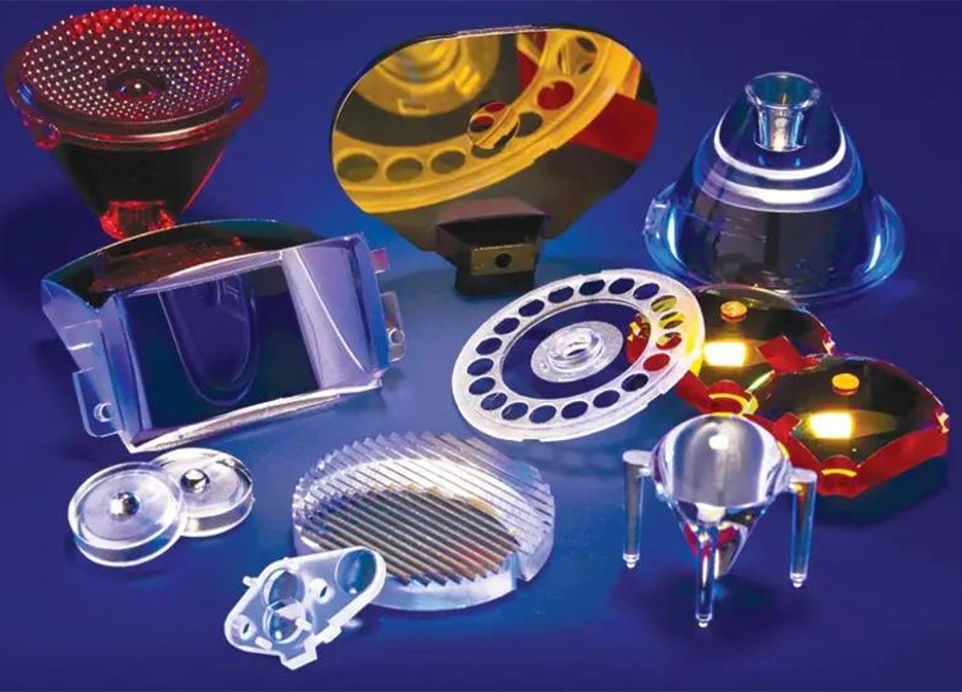 A variety of custom precision polymer optical components displayed on a blue background. The parts include lenses, reflectors, diffusers, and complex molded optical elements in various shapes and transparent or reflective finishes, demonstrating advanced polymer optics manufacturing capabilities.