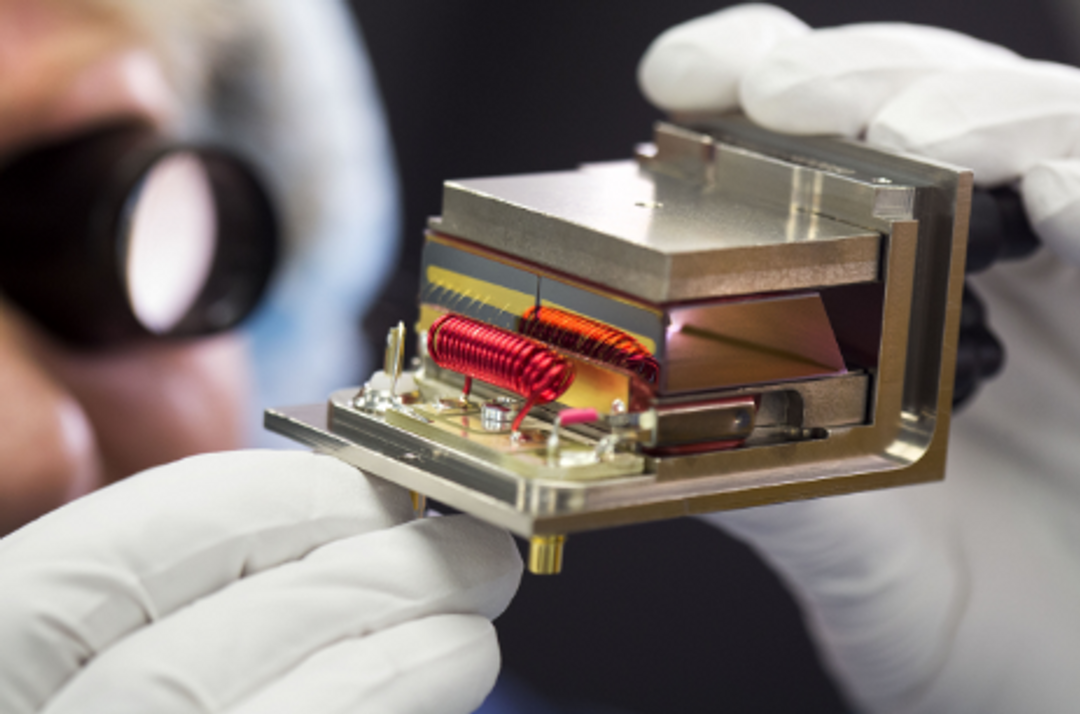 A close-up of a person wearing white gloves carefully holding a high-precision electronic or optical component. The device includes metallic parts, coils of red wire, and intricate internal structures, indicating it may be part of advanced scientific or aerospace equipment