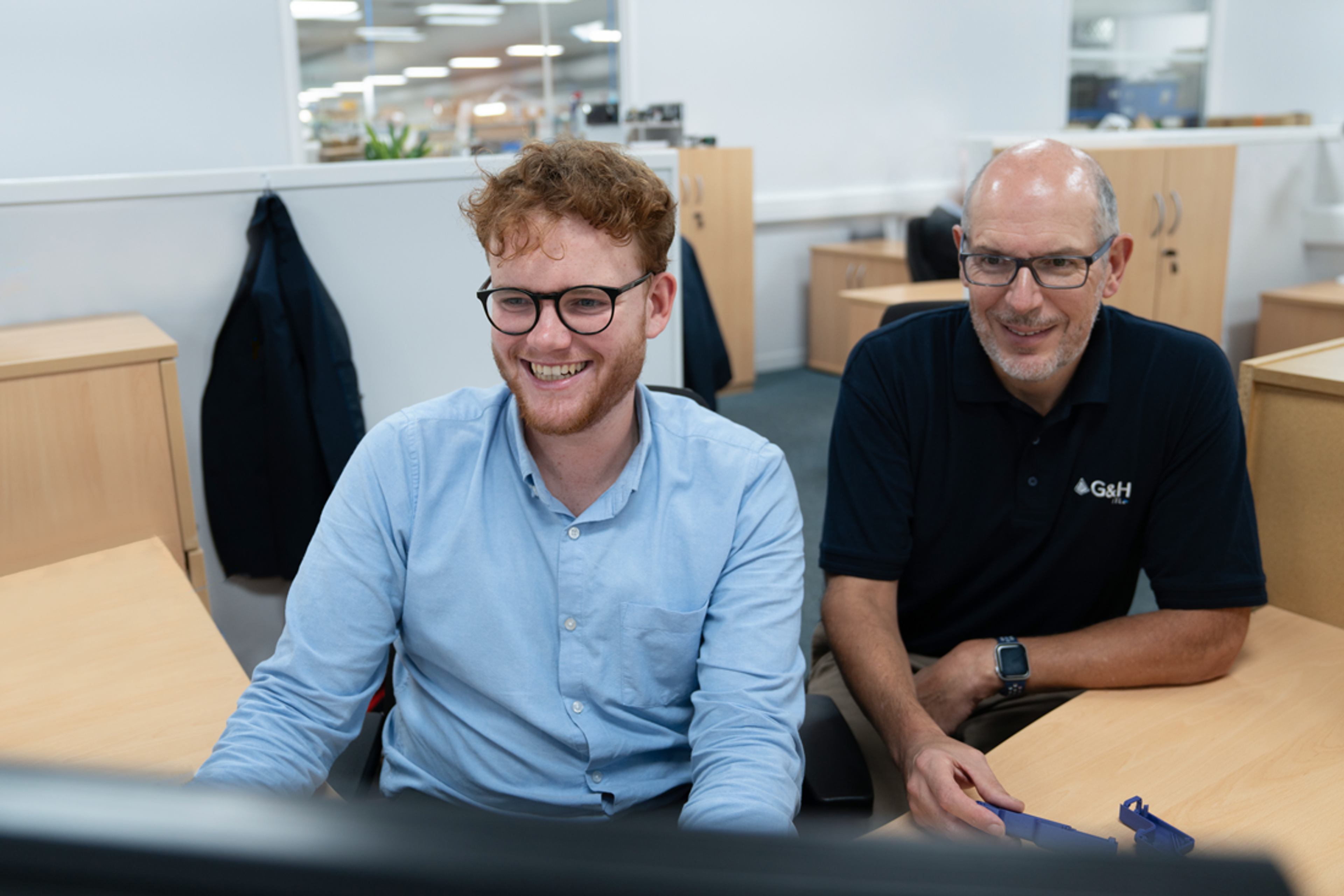Colleagues at ITL, Two men sit side by side at a desk in a bright, modern office, both smiling and looking at a computer screen. The man on the left, with curly red hair and glasses, wears a light blue shirt. The man on the right, wearing a black G&H polo shirt and glasses, holds a 3D-printed part. The background shows a tidy workspace with light wooden furniture and an open office layout