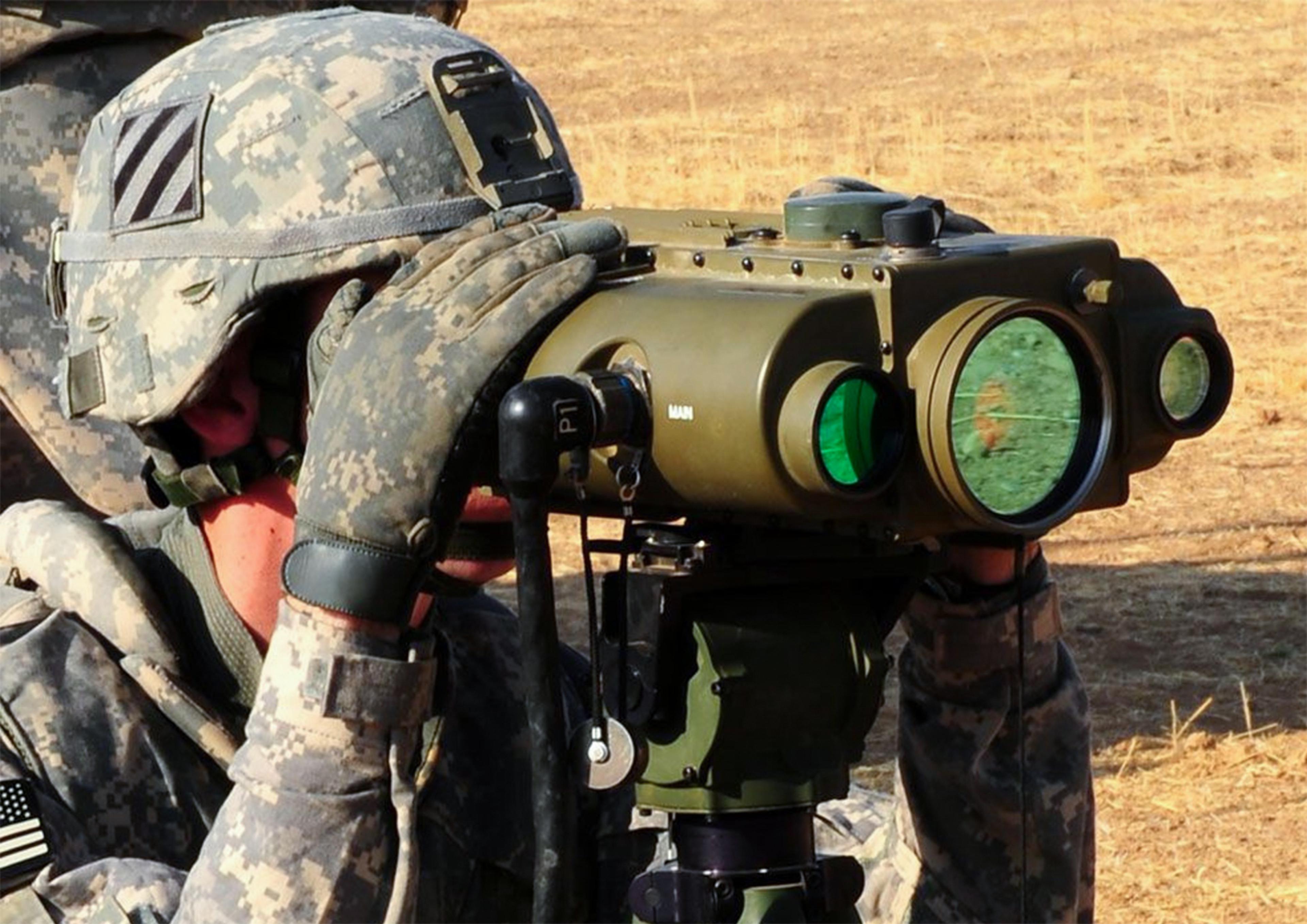 Soldier in digital camouflage operates a tripod-mounted target designation and laser rangefinding sight, looking through a large green-tinted optical lens in a dry, open field.