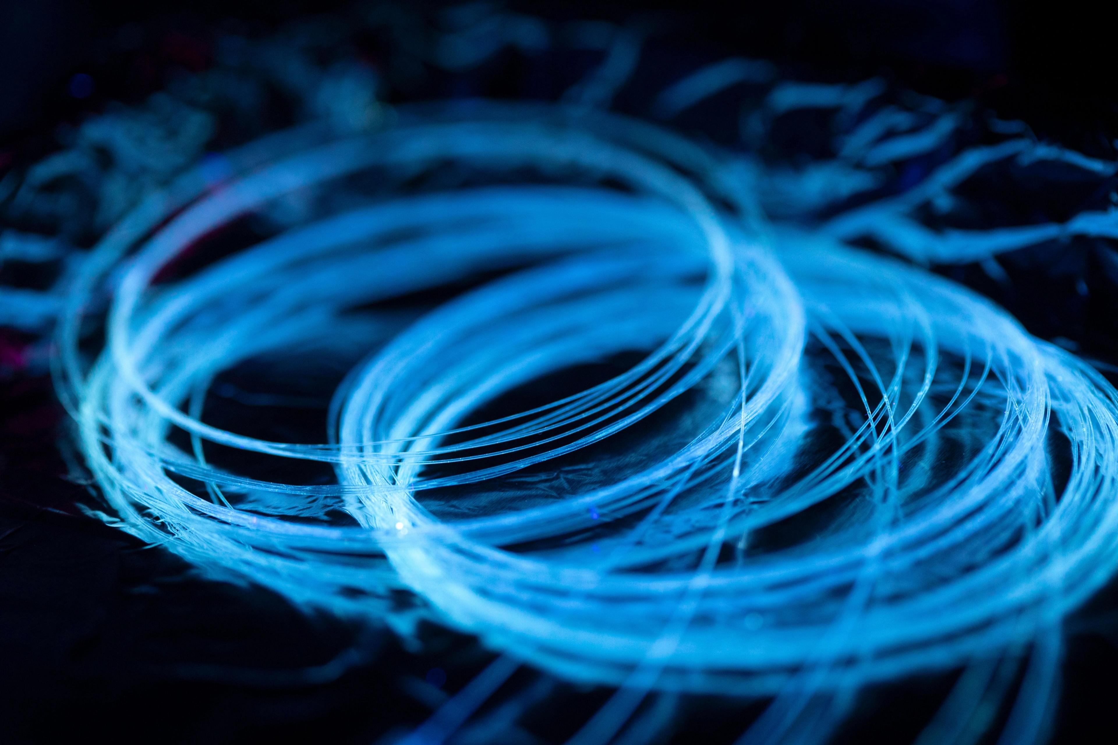 Close-up image of glowing optical fibers illuminated under UV light. The fibers form circular loops and emit a vibrant blue light against a dark background, creating a dynamic and abstract visual effect.