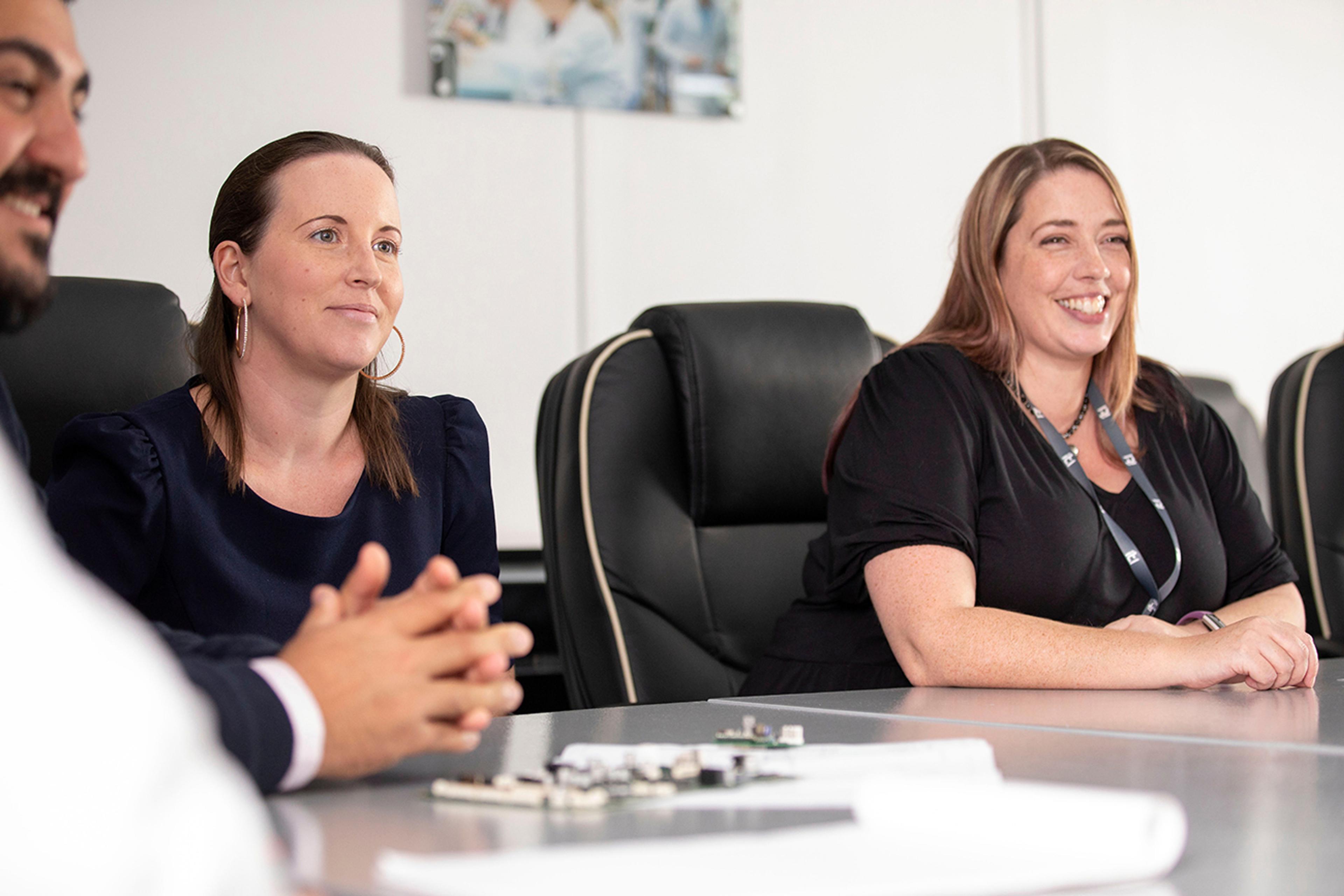 Three professionals are seated around a conference table engaged in a meeting. Two women in the foreground are smiling and appear to be listening attentively. They are seated in black office chairs, and documents and circuit boards are visible on the table. The setting is a modern, well-lit office environment.