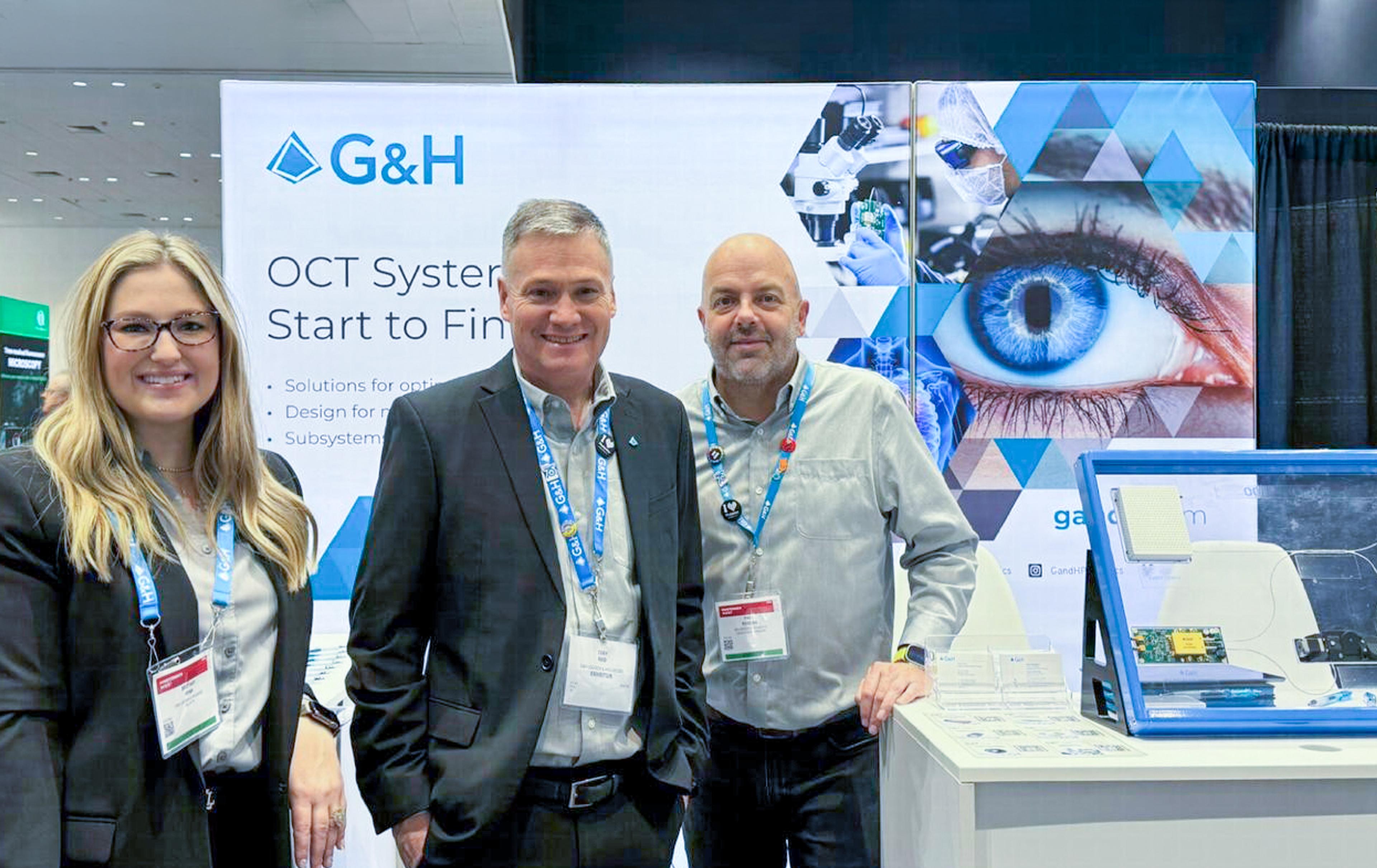 Three G&H team members at the Photonics West BiOS 2024 stand, alongside an OCT systems banner and a display of optical components.