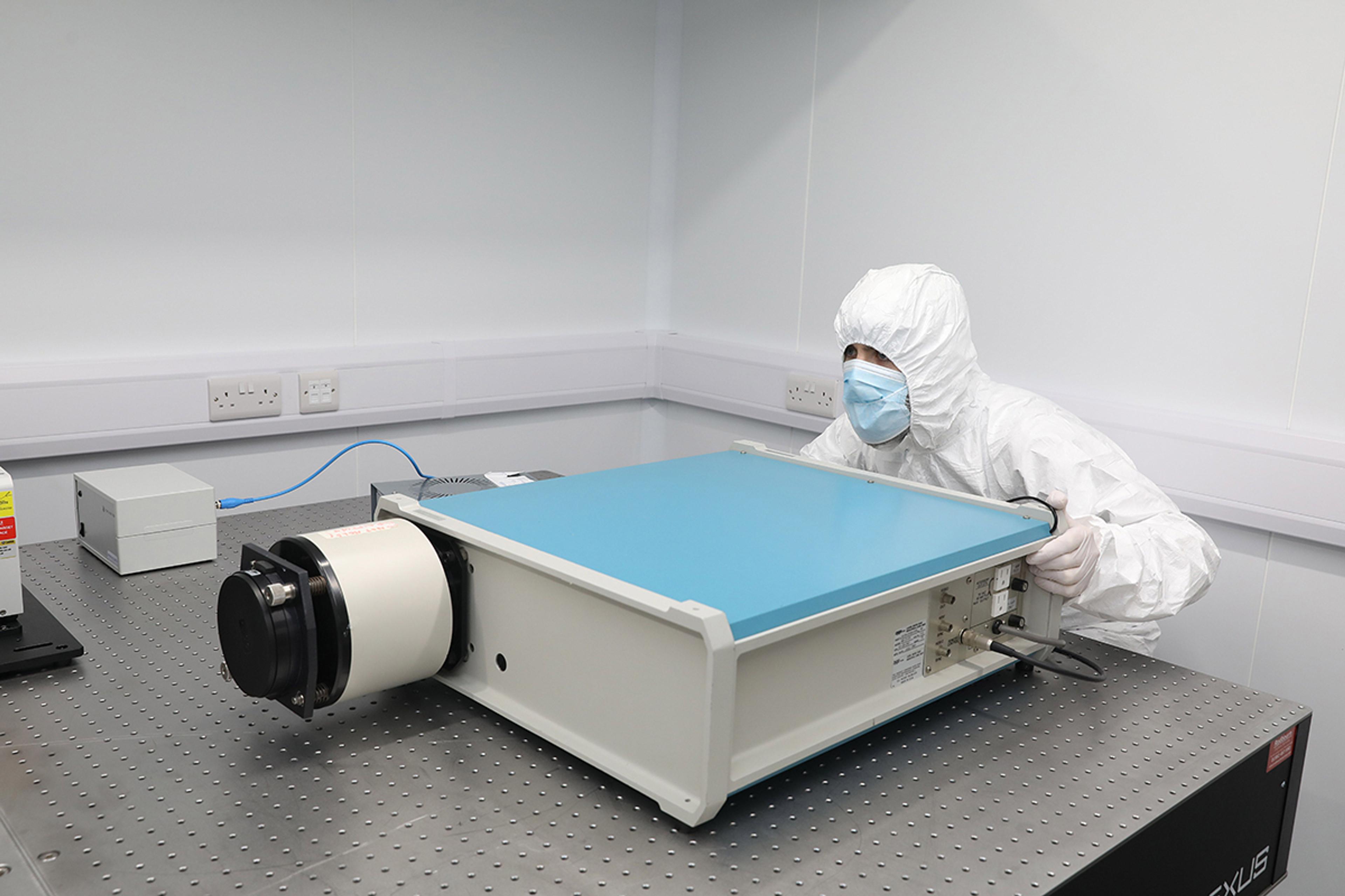 Scientist in full cleanroom attire, including hood, gloves, and face mask, positioning a large optical device with a blue top on an anti-vibration optical bench in a sterile laboratory environment.