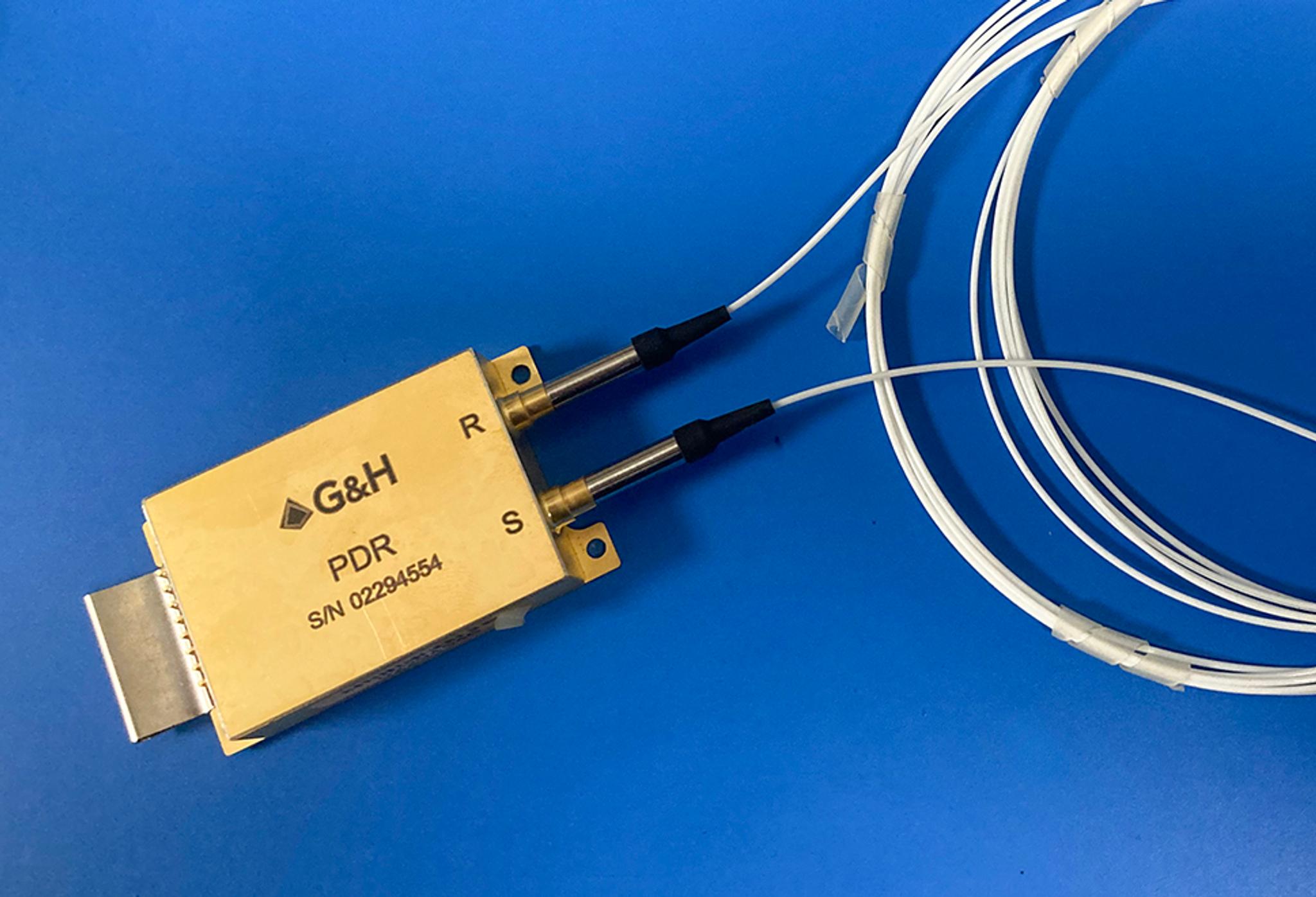 A gold-colored G&H PDR (Polarization Dependent Reflector) module with serial number 02294554, featuring two labeled ports ("R" and "S") connected to white optical fibers, coiled and secured with tape, resting on a blue surface.