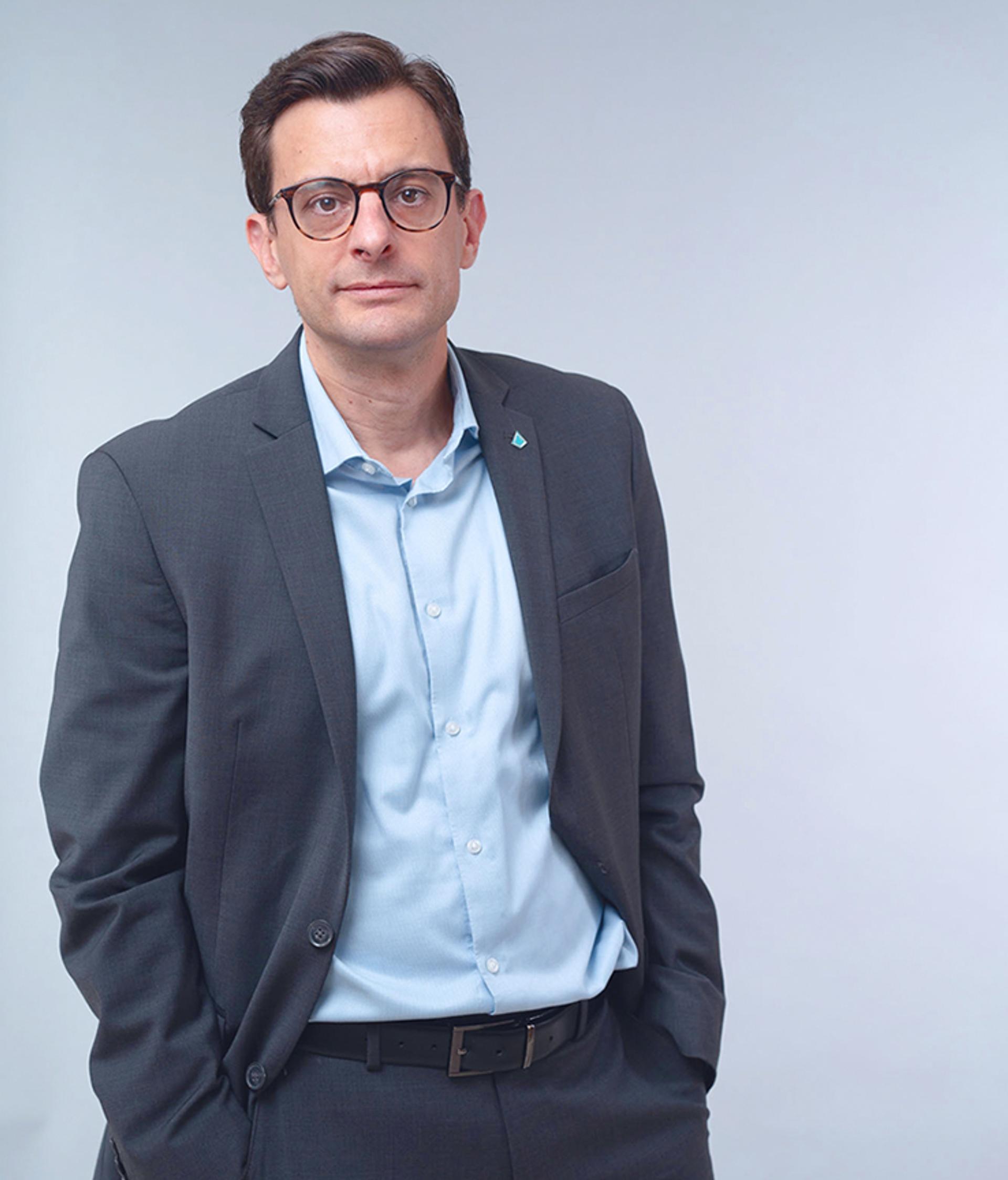 Portrait of a man wearing glasses, a dark grey suit and a light blue shirt, standing with his hands in his pockets against a pale blue background.