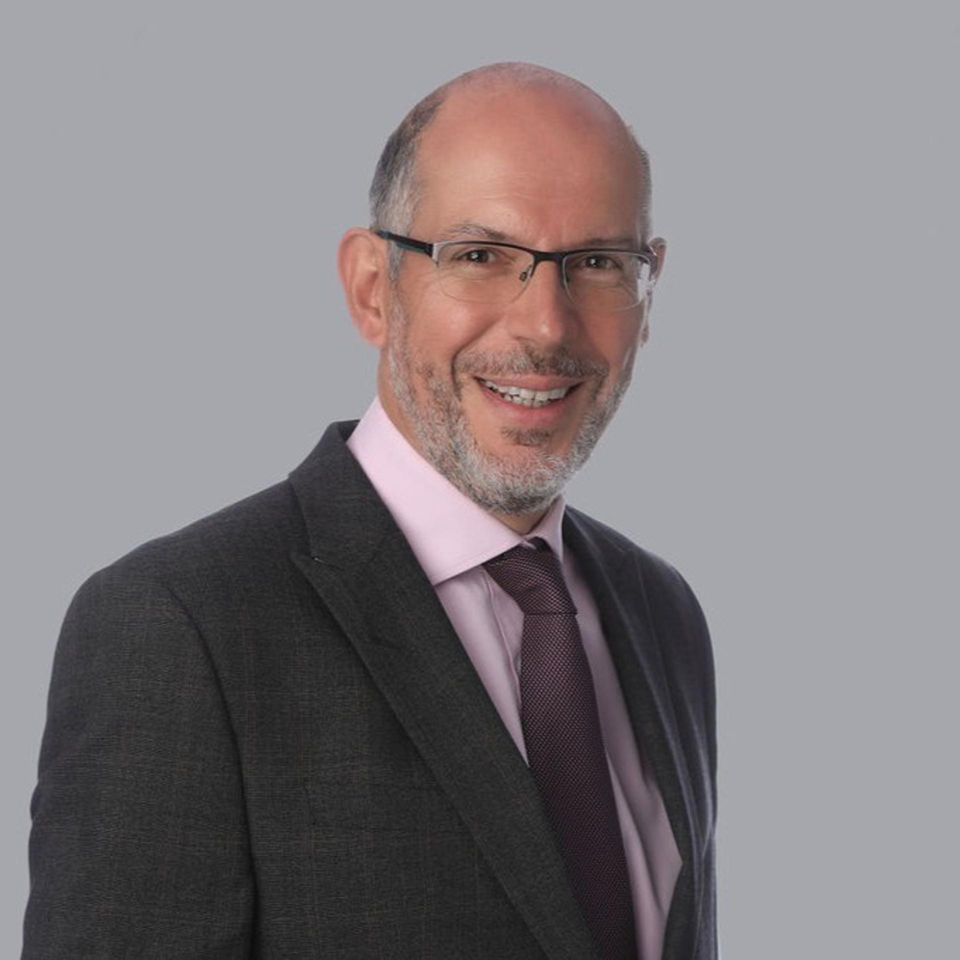 A middle-aged man with a bald head and neatly trimmed grey beard is smiling at the camera. He is wearing glasses, a dark grey suit, a light pink shirt, and a textured dark purple tie. The background is a plain light grey.