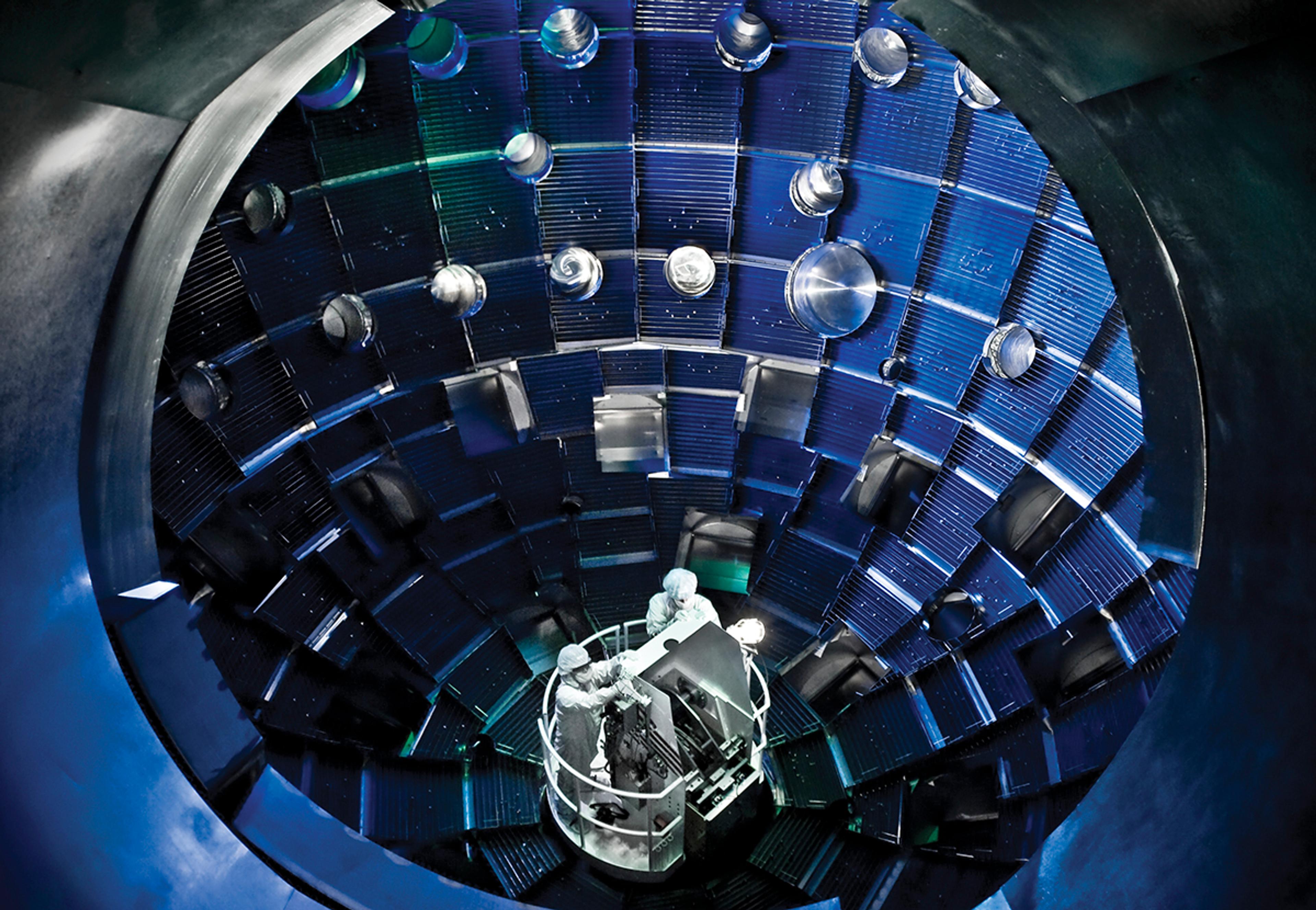Engineers working inside the spherical target chamber at the National Ignition Facility, surrounded by layered diagnostic and laser entry ports used in fusion experiments.