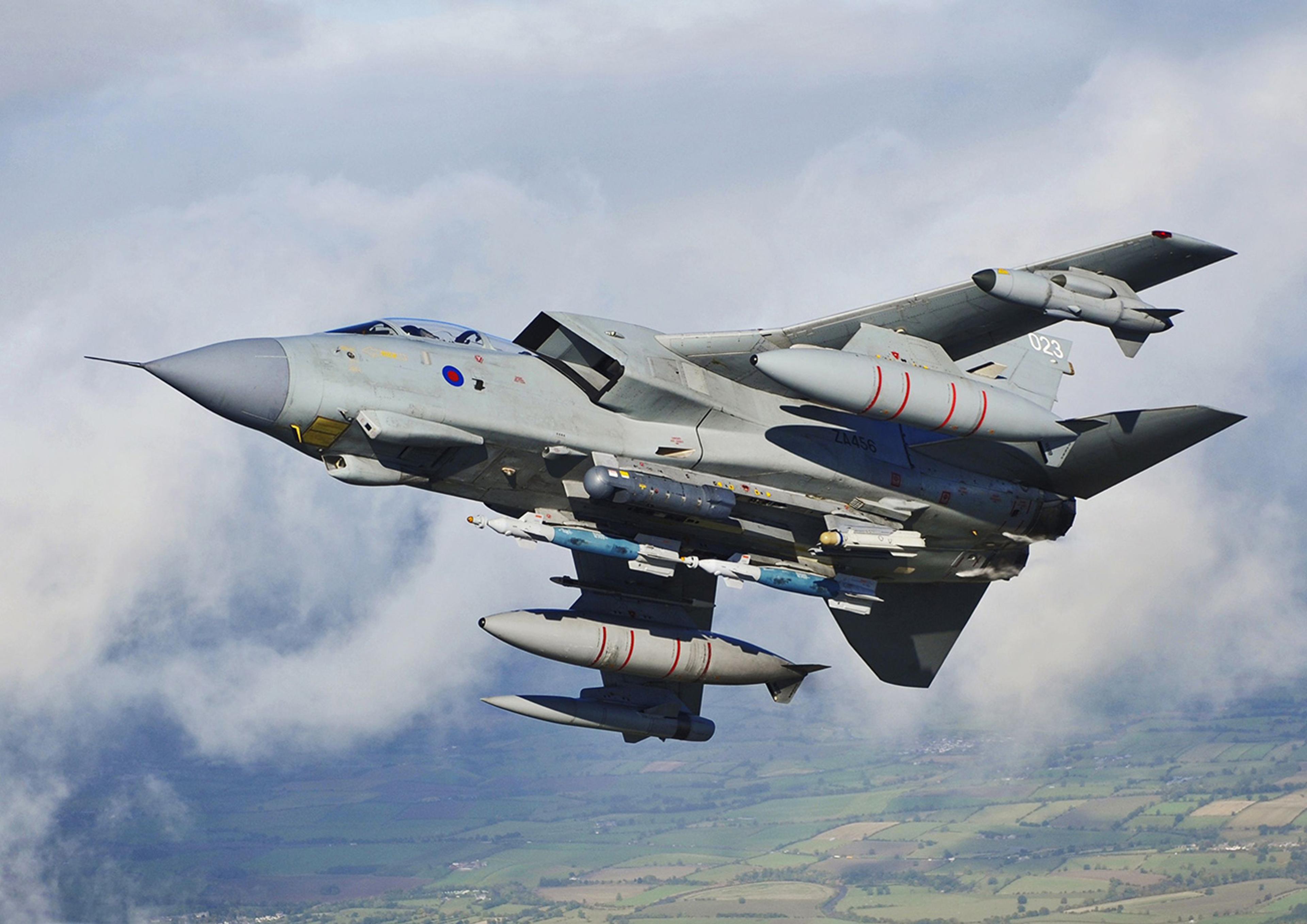 A military fighter jet in flight above a patchwork of fields and countryside, with clouds surrounding it. The aircraft is equipped with multiple weapons and fuel tanks under its wings, displaying a grey camouflage paint scheme and marked with identification numbers and insignia.