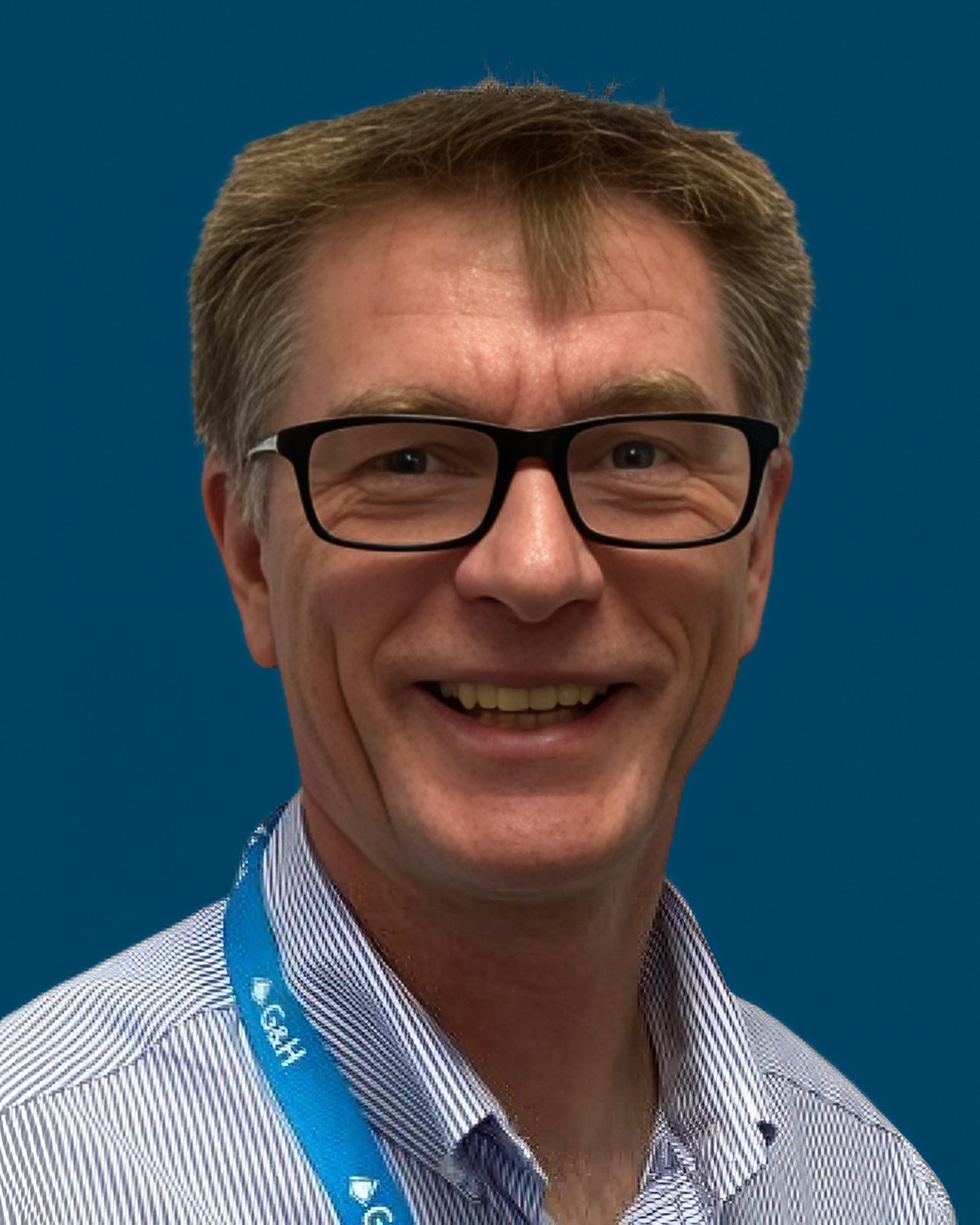 Head-and-shoulders portrait of a smiling man wearing black-framed glasses and a patterned shirt, photographed against a blue background.
