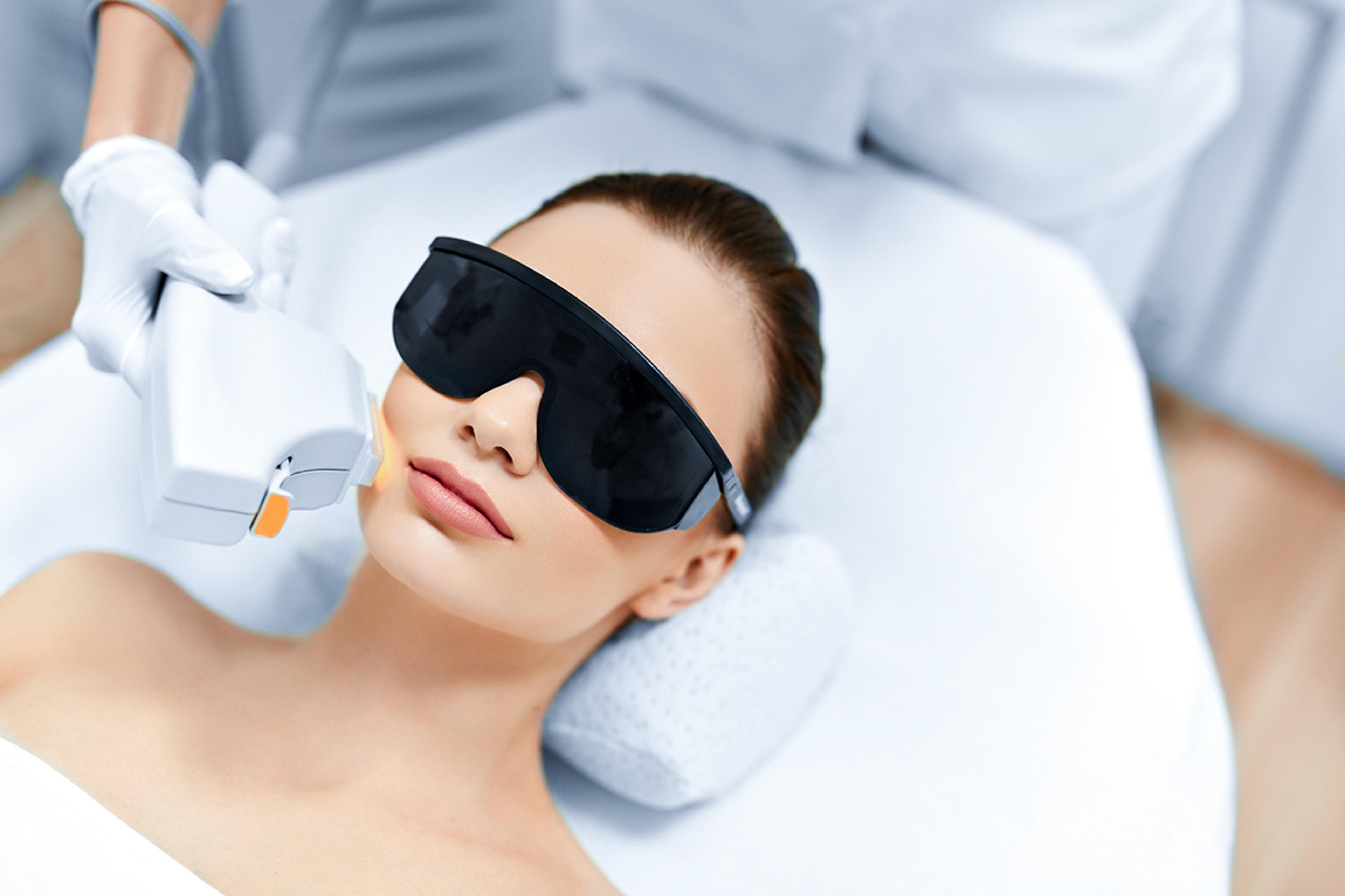 Aesthetic laser - A woman lies on a treatment bed wearing protective black goggles while undergoing a cosmetic procedure. A practitioner, dressed in white and wearing gloves, applies a handheld laser or intense pulsed light (IPL) device to the woman's face in a clinical or spa setting