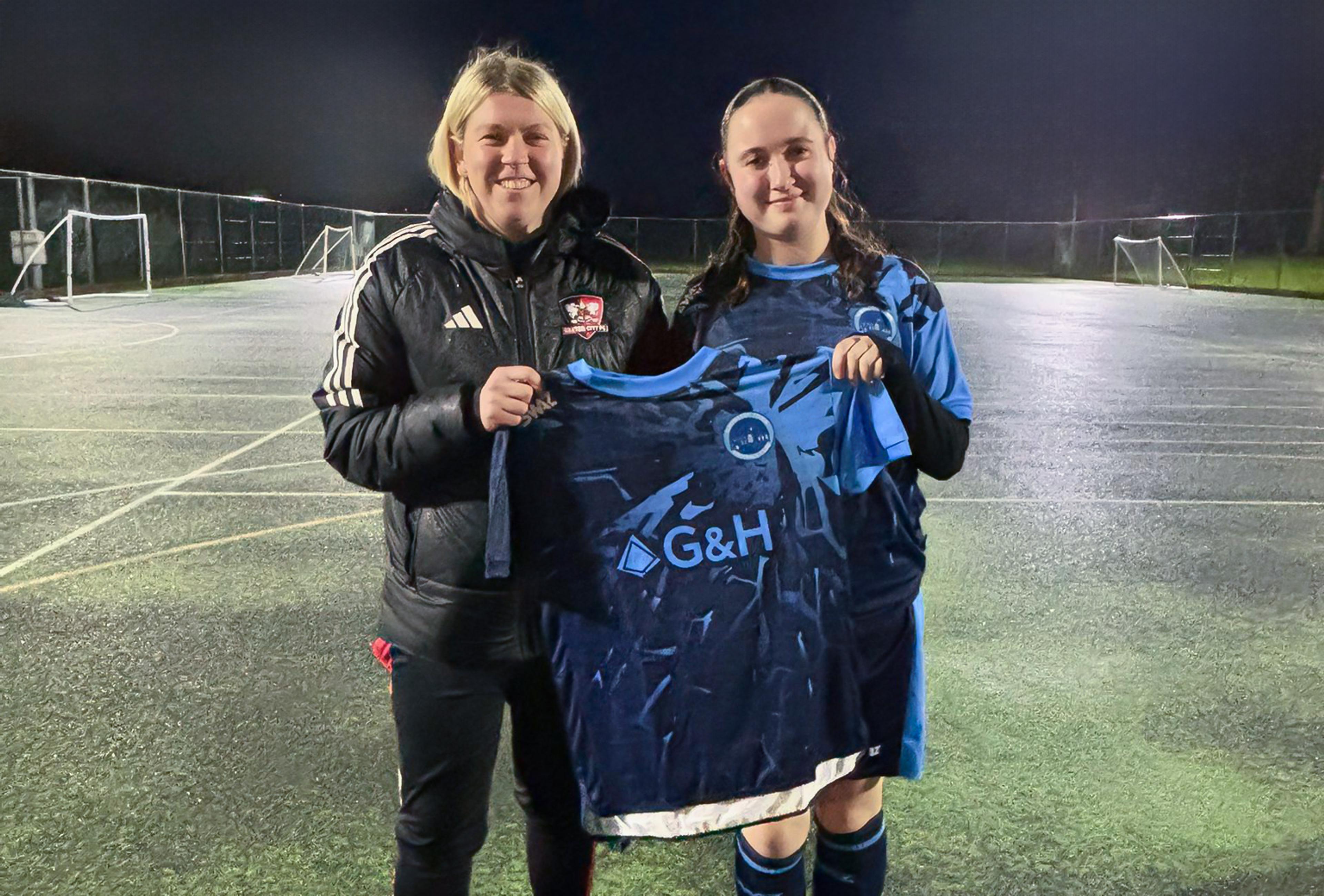 Two women stand on a floodlit outdoor football pitch at night, smiling and holding up a blue G&H-branded team shirt.