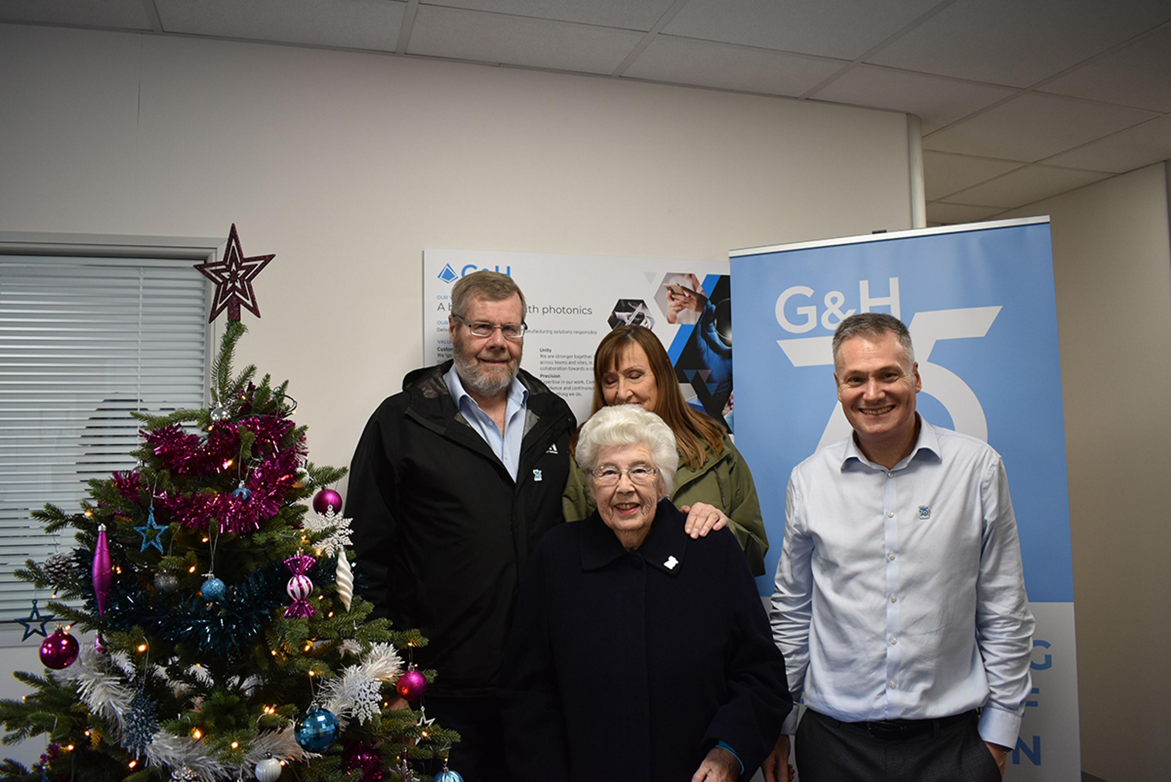 Norma Knapp (Centre) & family with Toby Reid (Right), Product Management Director, G&H Torquay. A group of five people pose together indoors next to a decorated Christmas tree. The elderly woman in front is smiling, dressed in a dark coat, and surrounded by four other adults—two men and two women—who are also smiling warmly. Behind them are banners and posters displaying the G&H logo, with one banner prominently showing “75” in celebration of the company’s anniversary. The setting suggests a festive event or company celebration during the holiday season.