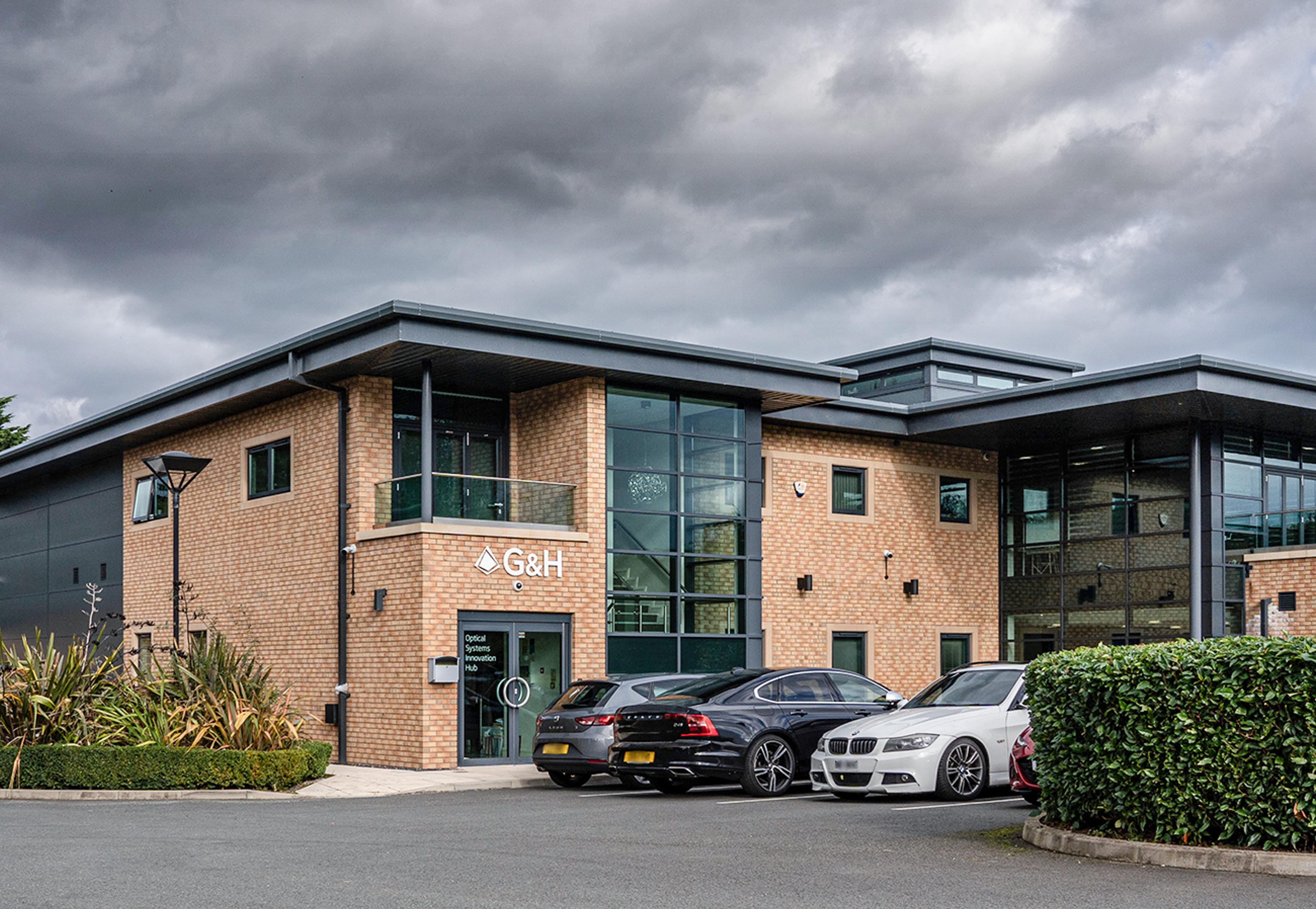 Exterior of the G&H Optical Systems Innovation Hub in St Asaph, showing the modern brick-and-glass facility with G&H signage and visitor parking.