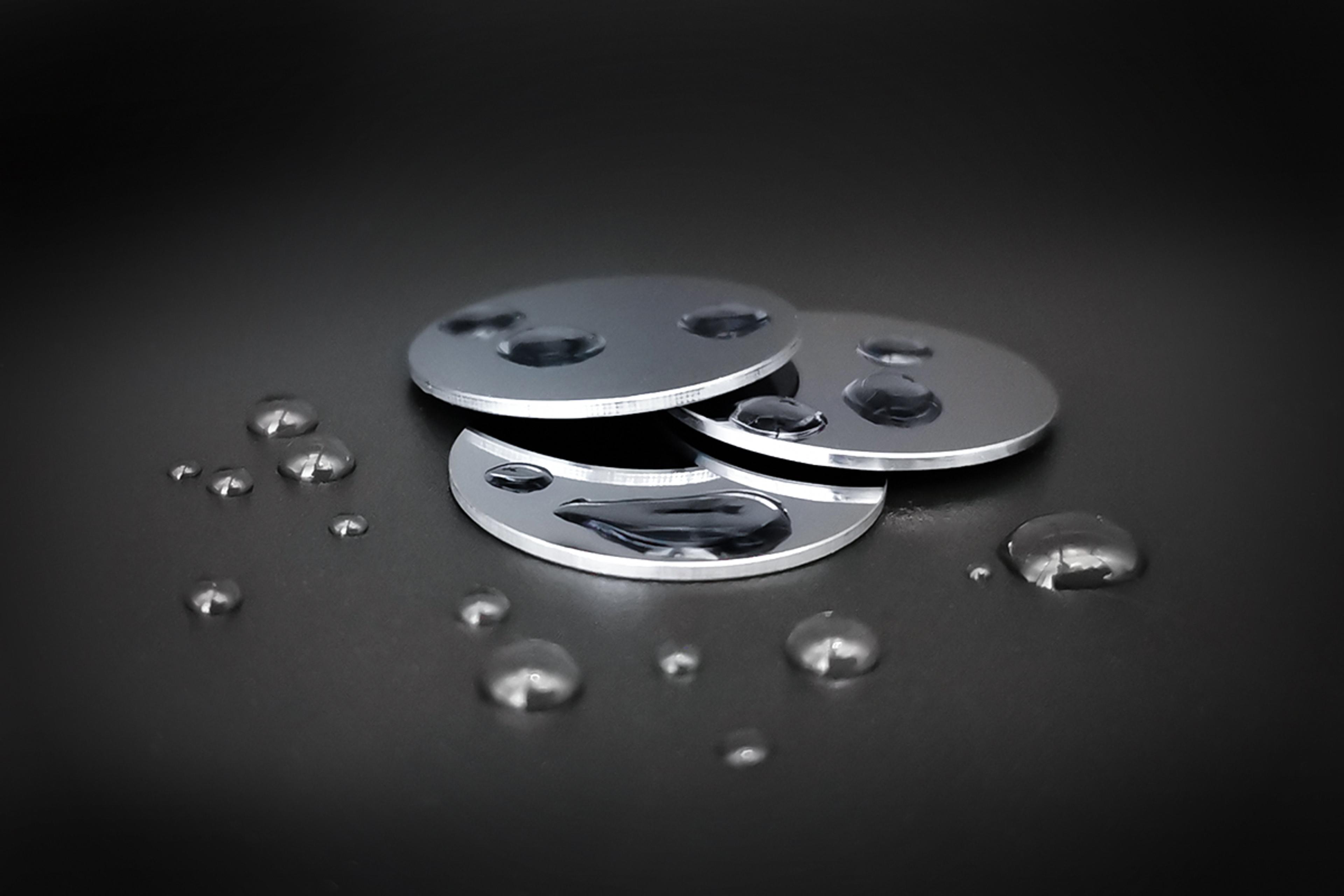A stack of polished circular Germanium wafers with water droplets on their surfaces, displayed against a dark background. The droplets highlight the hydrophobic or coated surface finish, suggesting precision fabrication for electronic or IR sensing applications.
