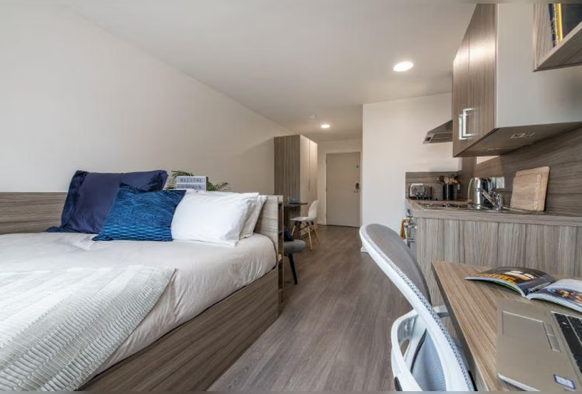 Studio apartment interior showing a bed with navy and teal cushions, a hallway leading to a kitchenette with wood cabinets, and a desk with laptop in the foreground.