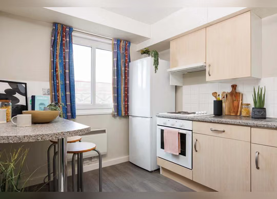 Compact kitchen with light wood cabinets, a white fridge-freezer and oven, patterned curtains, a small breakfast bar with stools, and a granite-effect countertop.