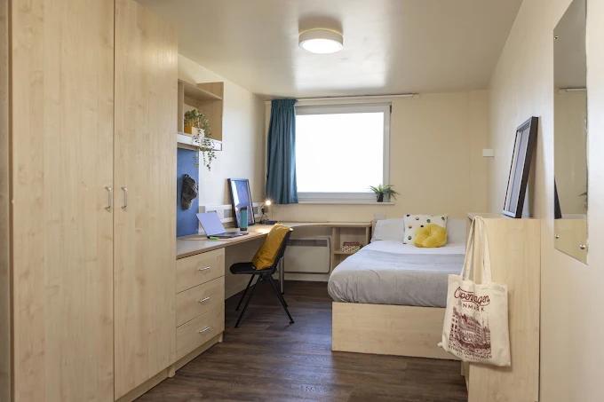 Bedroom with light wood wardrobes and desk, a laptop and monitor setup, teal curtains, a single bed with yellow cushion, and a tote bag hanging on the corner.