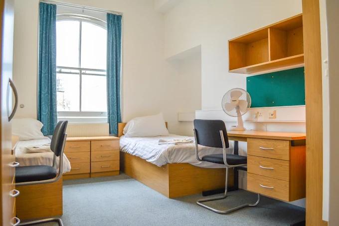 Twin bedroom with two single beds, wood chests of drawers, a desk with a green pinboard and desk fan, black chairs, and teal curtains on an arched window.
