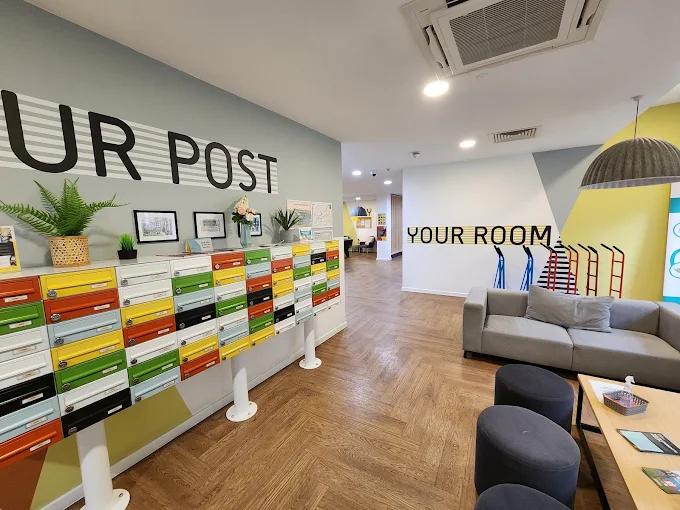 Building common area with a wall of colorful mailboxes beneath a "Your Post" sign, and a grey sofa and seating area near a "Your Room" wall graphic.