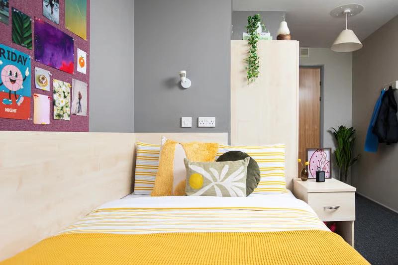 Student bedroom with a yellow-striped bed, patterned throw pillows, a wood headboard, a corkboard collage on the wall, and a wardrobe with hanging plants on top.