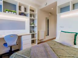 Student bedroom with a built-in white desk and shelving unit, blue desk chair, and a bed with patterned bedding and green accent cushion.