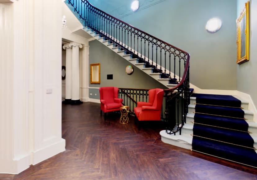 Elegant entrance hall with a grand staircase, dark wood flooring, decorative columns, and two red armchairs in a spacious reception area.