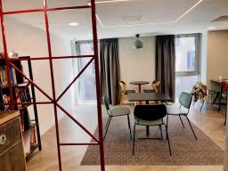 Communal dining area with a dark wood table and mismatched chairs, near a red-framed bookshelf and curtained windows.