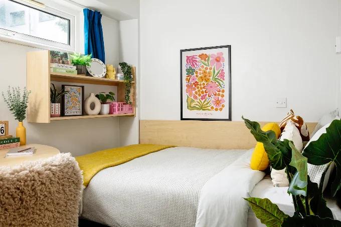 Bedroom with a wood headboard, floral wall art, a wall-mounted shelf with plants and decor, blue curtains, and a bed dressed with white and mustard-yellow bedding