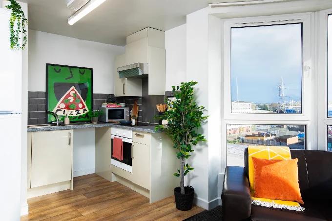Studio kitchenette with cream cabinets, a pizza-themed art print, and a window with harbor views beside a brown sofa with yellow and orange cushions.