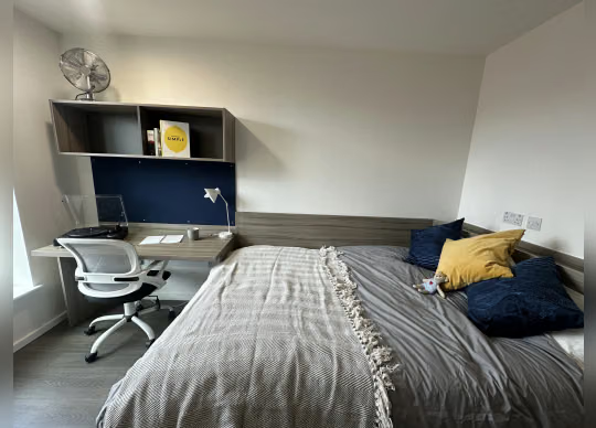 Bedroom with a grey and navy color scheme, a desk with an oscillating fan and wall-mounted shelving, and a bed with grey throw and mustard and navy cushions.