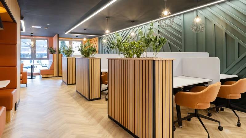 Co-working/study nook with wood-slat partition booths, potted plants, mustard-colored chairs, and patterned green wall panels.
