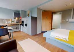 Studio apartment with a kitchenette on the left, wooden flooring, and a bed with yellow and white bedding on the right, plus a wooden door leading to another room.