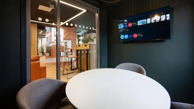 Private meeting or cinema room with a round white table, grey armchairs, and a wall-mounted TV displaying streaming app icons.