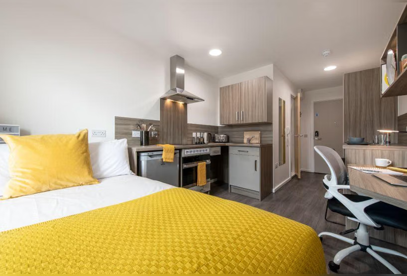 Studio apartment with a bed featuring a yellow bedspread and pillow, an integrated kitchenette with wood cabinets, and a desk and chair near the entrance door.