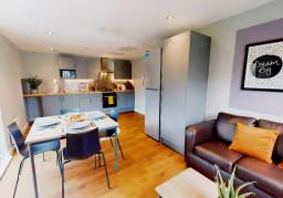 Open-plan studio kitchen and living space with a dining table and chairs, kitchen units, a brown sofa with orange cushion, and a framed wall print reading "Dream."
