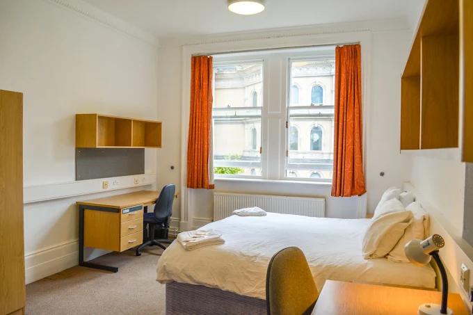 Single bedroom with a wood desk and shelving unit, orange curtains, a large sash window overlooking a period building, and a bed with white linens.
