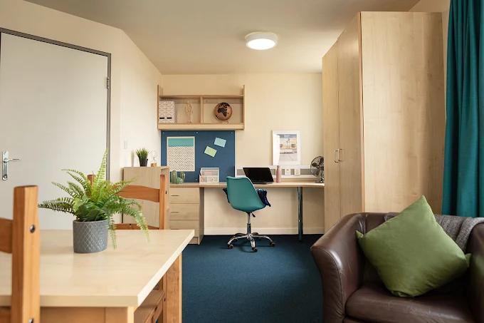 Bedroom corner with a wooden desk and shelving unit, a blue pinboard, teal office chair, wardrobe, dark green curtains, and a brown leather sofa with a green cushion.