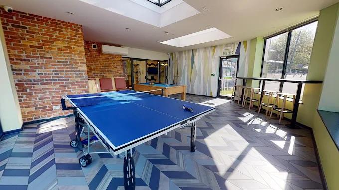 Bright communal games room with a blue table tennis table, exposed brick wall, skylight, pool table in the background, and tall windows overlooking a courtyard.