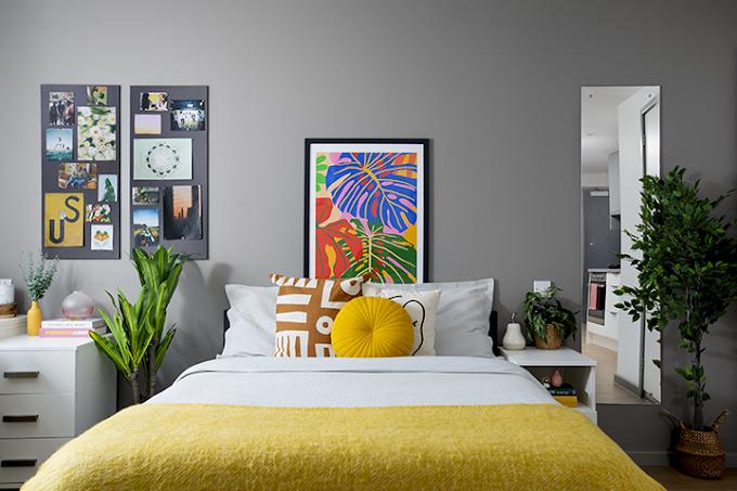 Bedroom with a grey accent wall, colorful botanical wall art, a bed with yellow throw and patterned cushions, white bedside units with plants, and a mirror reflecting a kitchenette beyond.