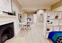 Modern studio apartment with a double bed, study desk, shelving unit, fireplace, and open-plan living area featuring light wood flooring.