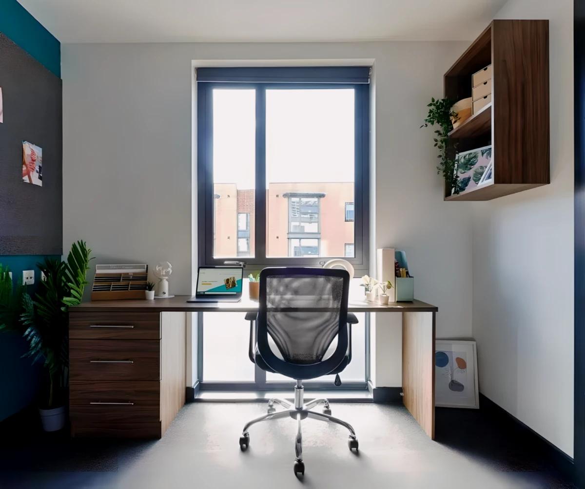 Study desk area in a student bedroom with a black office chair, wooden drawers, wall-mounted storage shelf with plants, positioned in front of a large window with a view of nearby buildings.