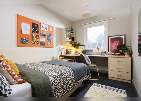 Bedroom with a patterned duvet and cushions, an orange pinboard with photos above the bed, a wooden desk with lamp and laptop, and a yellow patterned rug.