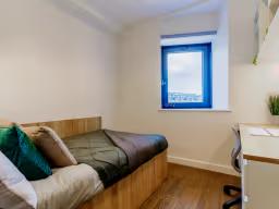 Compact student bedroom with a wooden-framed bed with green and cream cushions, a bright blue-framed window, and a desk visible in the corner.