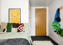 Bedroom corner showing a bed with patterned cushions, a wooden door, and a coat hook with a green bag hanging.